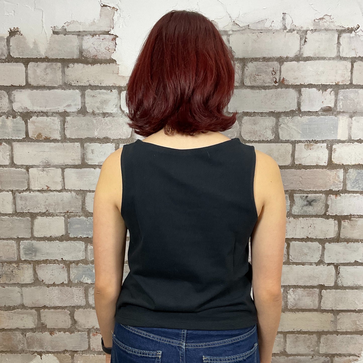 Person wearing a black sleeveless top and blue jeans against a brick wall. Upcycled fender guitar tshirt by unwantees, recycled tank tops, one of a kind