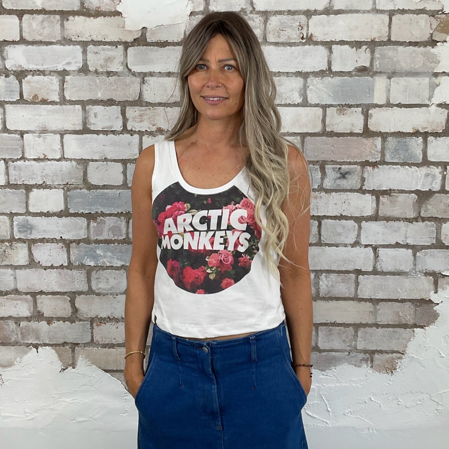 Woman wearing a white tank top with Arctic Monkeys logo in front of a textured brick wall. Arctic Monkeys Floral Tank Top, Vibrant coloured top by Arctic Monkeys Up Cycled recycled by Unwantees