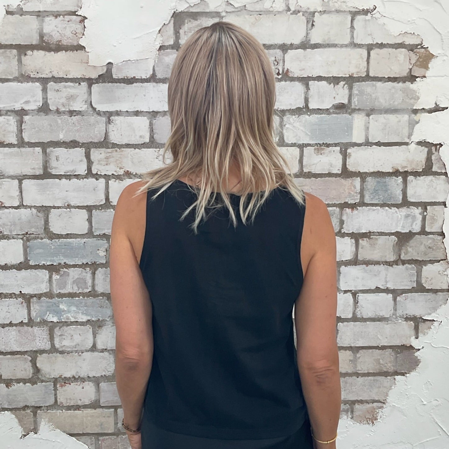 Person wearing a black sleeveless Unwantees tank top standing in front of a textured white brick wall.