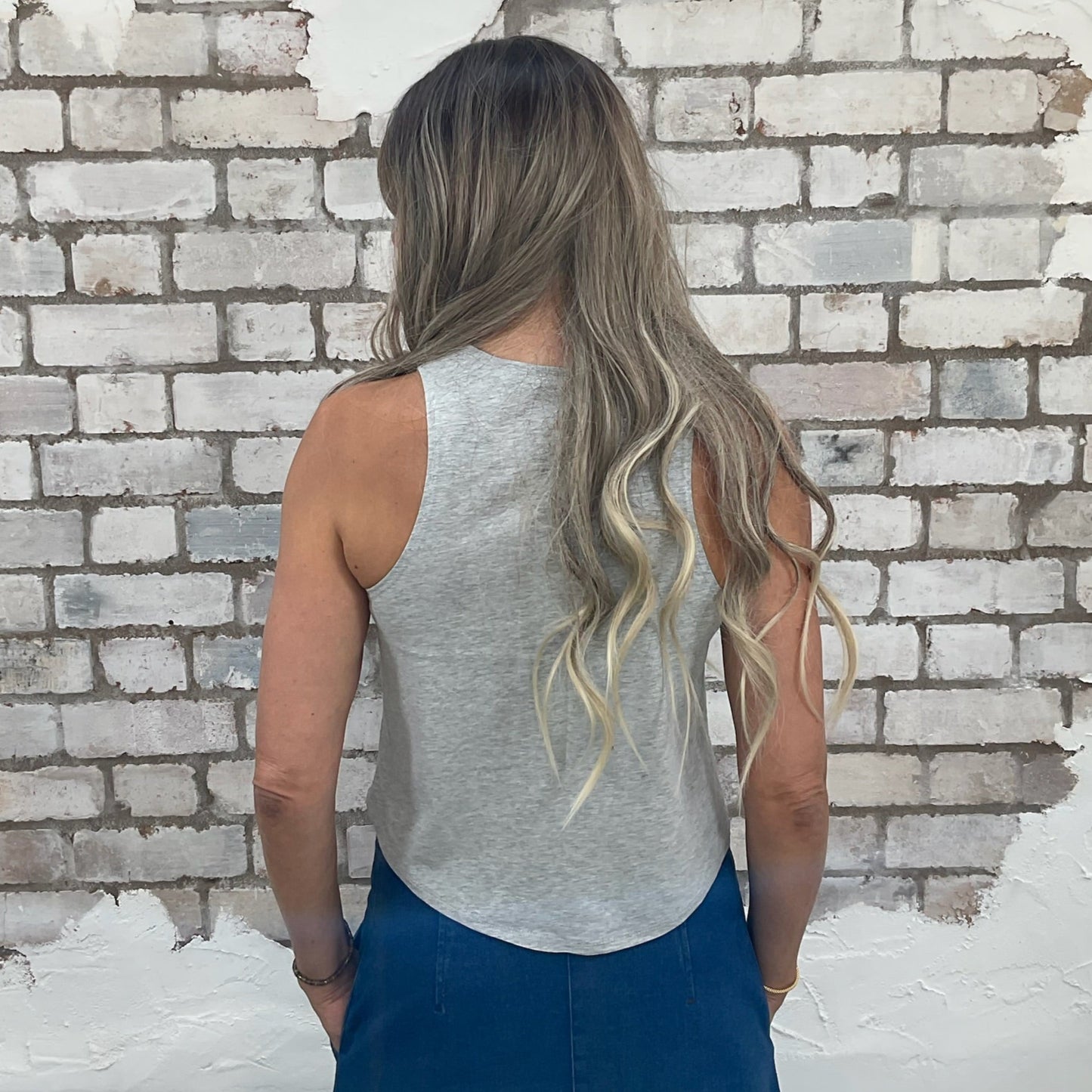 Woman with long blonde hair wearing a gray tank top and blue pants standing against a white brick wall. Grey Racerback Unwantee Tank top, vest top