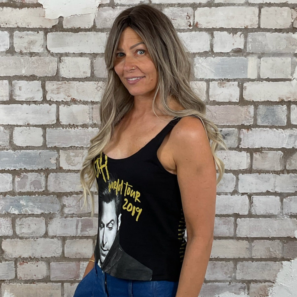 Woman wearing a black tank top with yellow text and blue pants standing in front of a textured white brick wall. Hugh Jackman low neck tank top, scoop neck tank, vest top, world tour 2019
