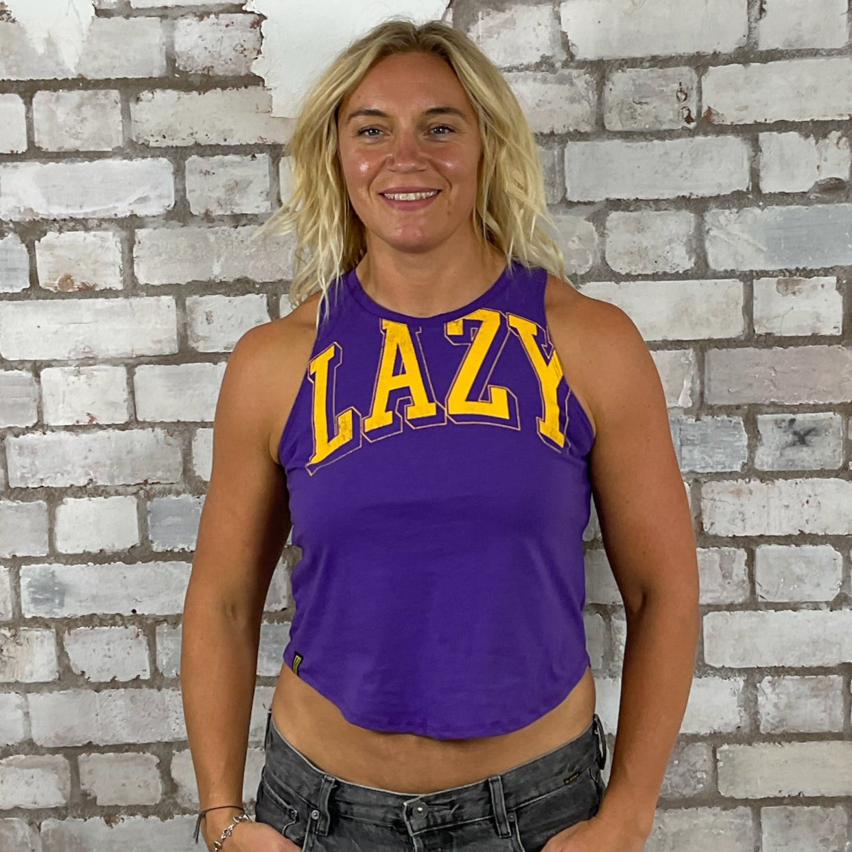 Person wearing a purple tank top with 'LAZY' text in front of a textured brick wall. Lazy Yellow print on purple tank top, vest top, tshirt up cycle by Unwantees