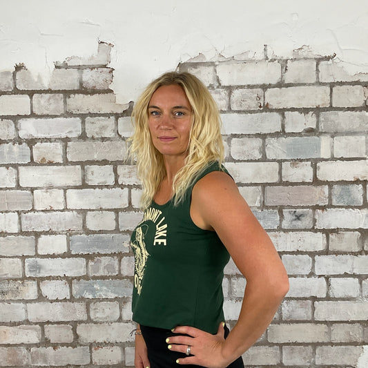 Woman wearing a green tank top with text against a white brick wall. Green Unwantee, Graan tank top, vest top, Piedmont lake, fishing print