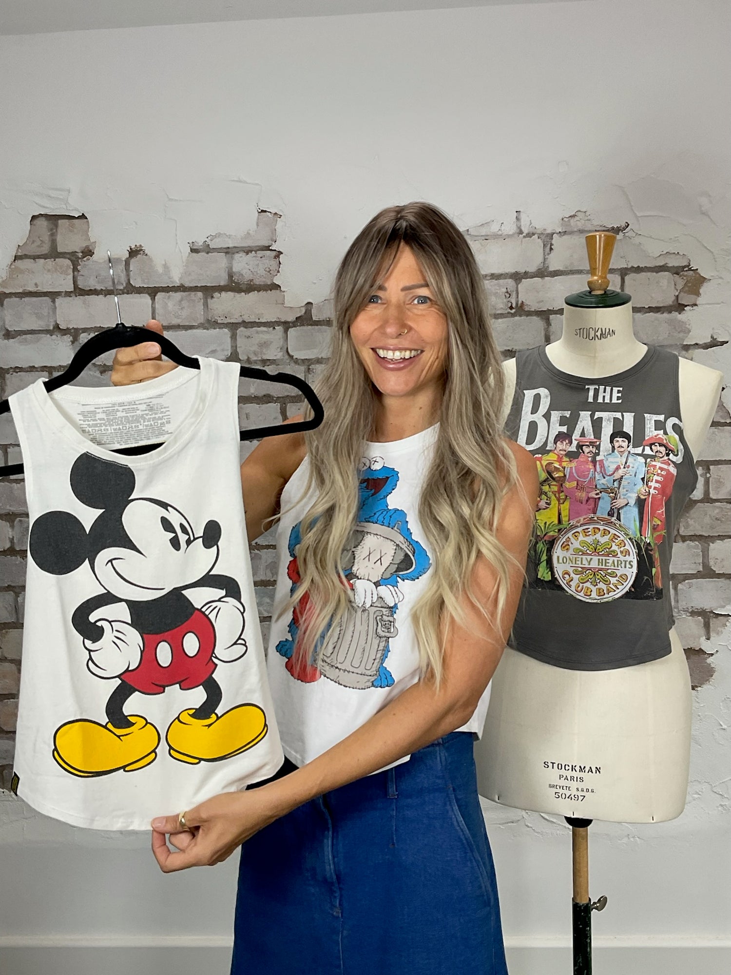 Owner Vinny smiling, holding a Mickey Mouse Unwantee, wearing an Elmo Unwantee, with a Beatles Unwantee on a mannequin behind her against a white brick wall