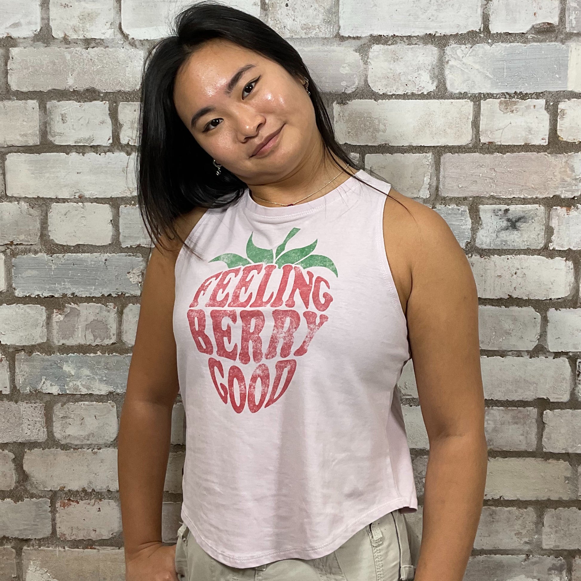Woman wearing a tank top with 'Feeling Berry Good' text against a brick wall.  Baby pink graphic tshirt upcycled by unwantees, tank top upcycled by unwantees, atleisurewear recycled clothing, sustainable clothing brand