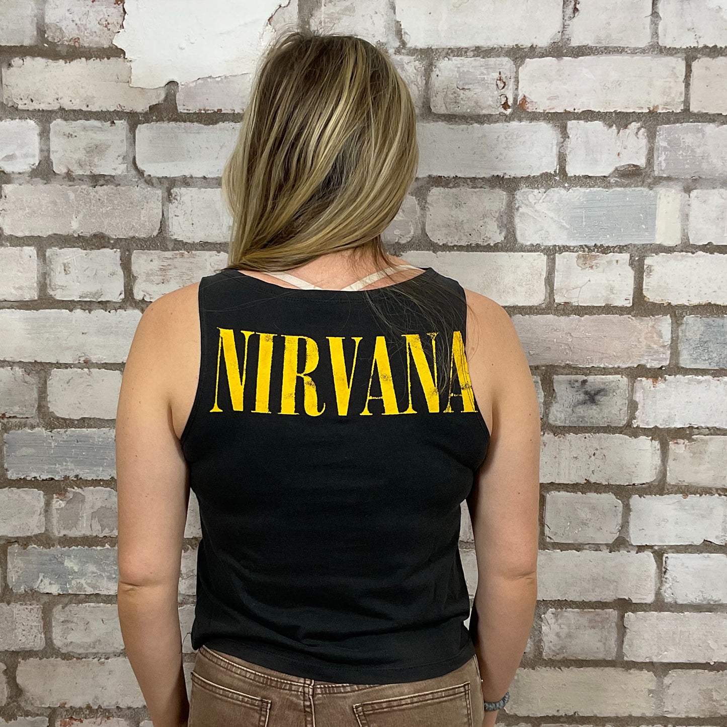 Person wearing a black tank top with 'NIRVANA' printed in yellow against a brick wall.  One-of-a-kind Nirvana Acid Face Yellow Print graphic Slash Neck Unwantee tank top close-up view, upcycled, sustainable, made in Britain