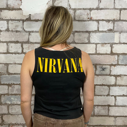 Person wearing a black tank top with 'NIRVANA' printed in yellow against a brick wall.  One-of-a-kind Nirvana Acid Face Yellow Print graphic Slash Neck Unwantee tank top close-up view, upcycled, sustainable, made in Britain