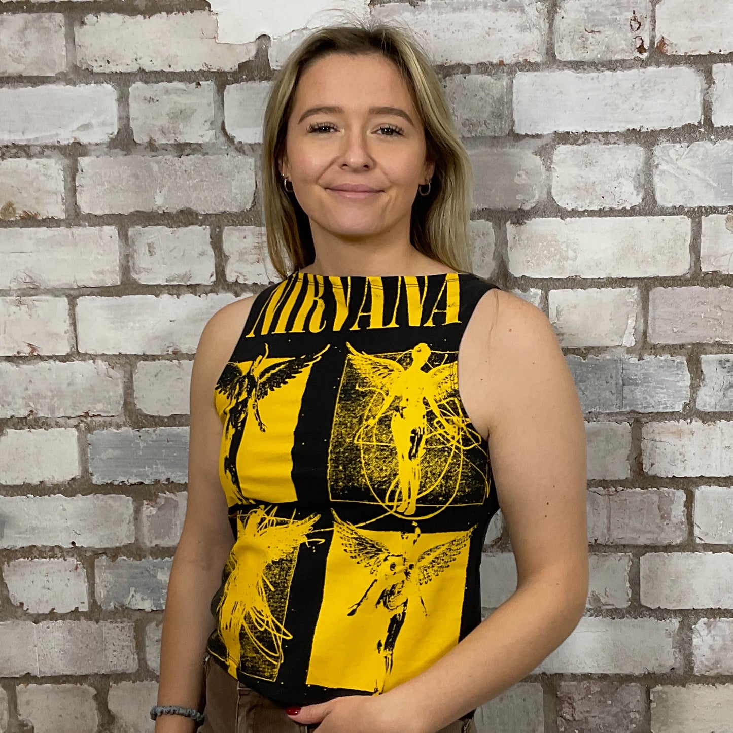 Woman wearing a black and yellow sleeveless top with fairy designs against a brick wall.  One-of-a-kind Nirvana Acid Face Yellow Print graphic Slash Neck Unwantee tank top close-up view, upcycled, sustainable, made in Britain