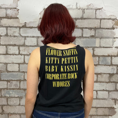 Person wearing a black tank top with yellow text against a brick wall.  One-of-a-kind Nirvana Acid Face Yellow Print graphic Slash Neck Unwantee tank top front view, upcycled, sustainable, made in Britain