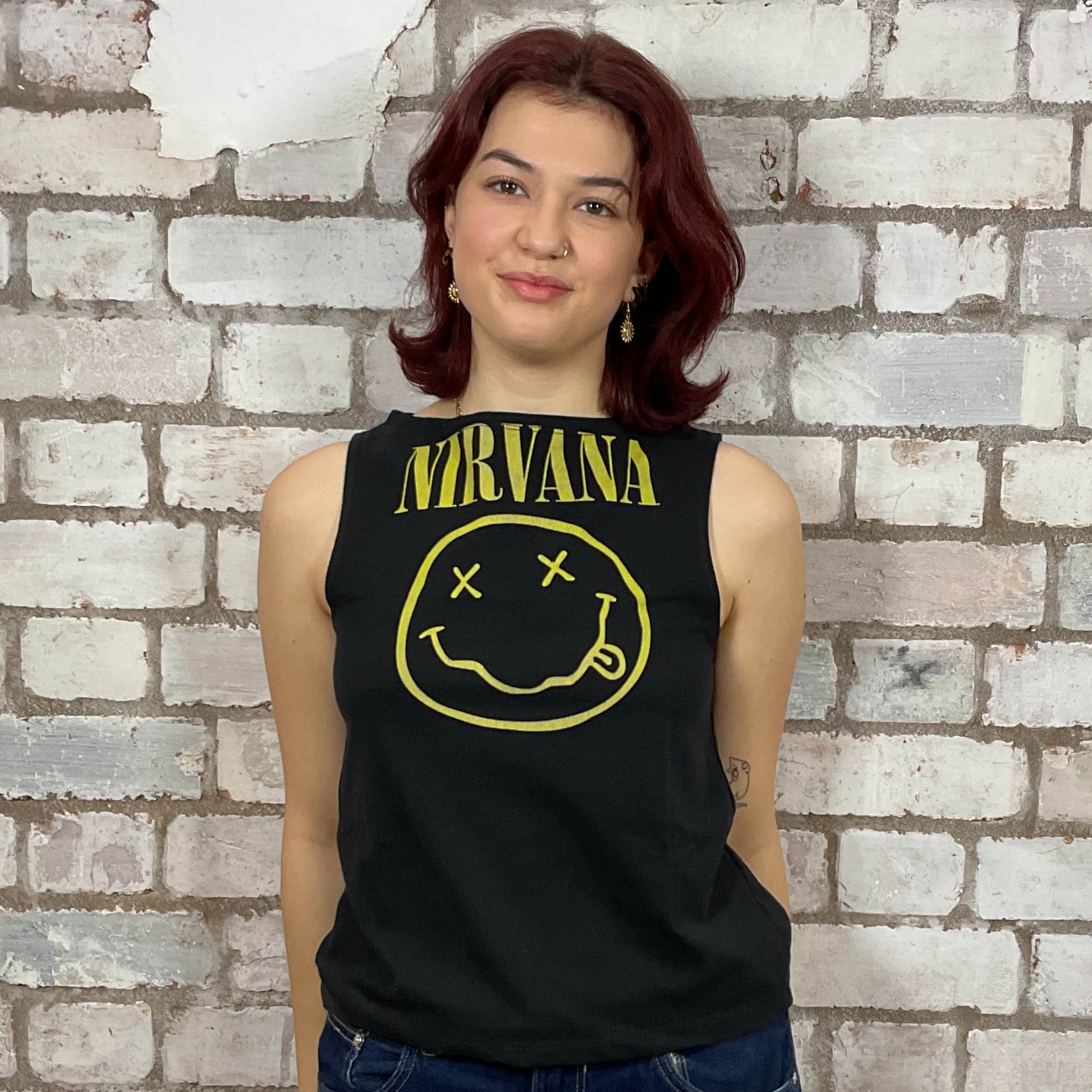 Person wearing a Nirvana tank top with smiley face design against a brick wall.