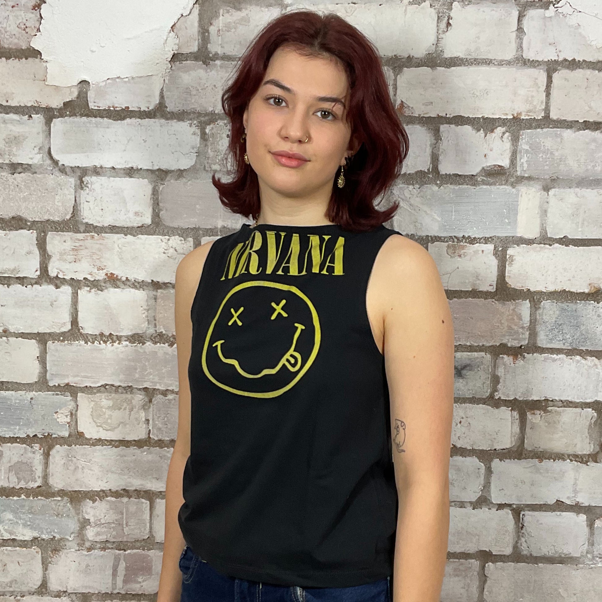 Person wearing a black tank top with Nirvana logo against a brick wall.  One-of-a-kind Nirvana Acid Face Yellow Print graphic Slash Neck Unwantee tank top front view, upcycled, sustainable, made in Britain