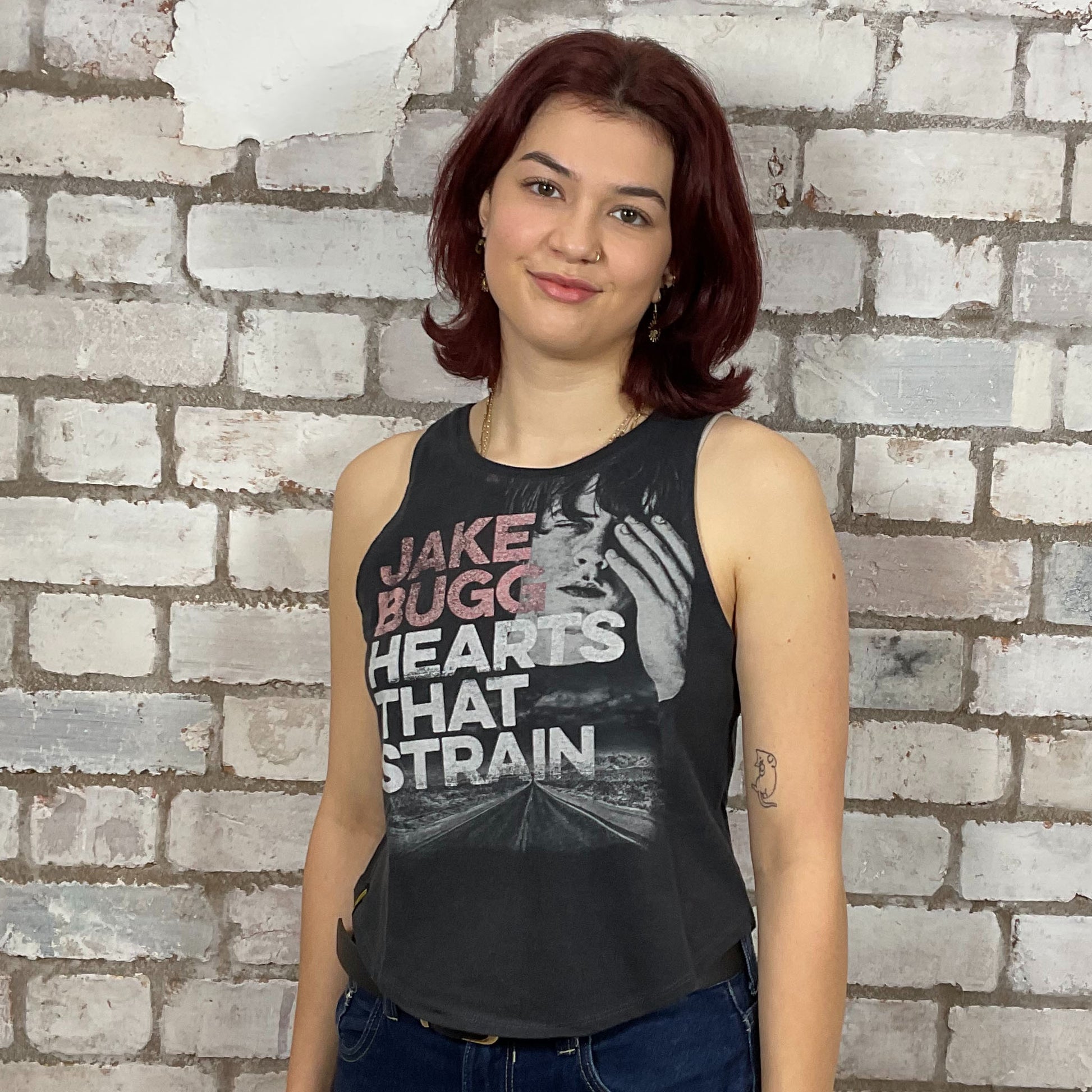 Person wearing a sleeveless shirt with 'Jake Bugg Hearts That Strain' text against a brick wall.  Jake Bugg Hearts that Strain Tour Tshirt upcycled by unwantees, recycled tank tops made in the uk