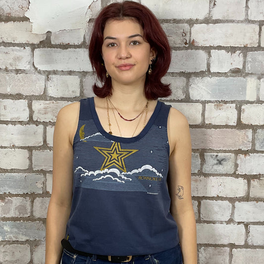 Woman wearing a blue tank top with a star design against a brick wall.  One-of-a-kind Roanoke VA Mystical Night Sky graphic Scoop Neck Unwantee tank top front view, upcycled, sustainable, made in Britain