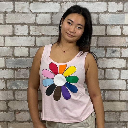 Person wearing a tank top with a colorful flower design against a brick wall.  One-of-a-kind Rainbow Flower graphic Scoop Neck Unwantee tank top front view, upcycled, sustainable, made in Britain