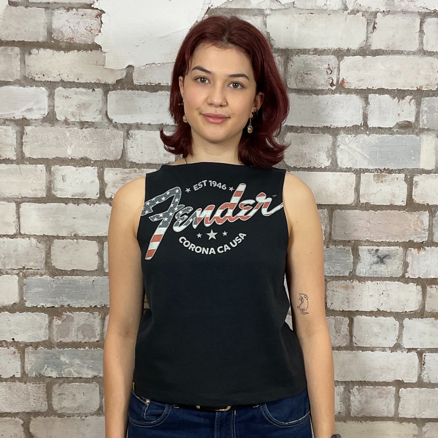 Woman wearing a black sleeveless shirt with 'Fender' logo against a brick wall.  Upcycled fender guitar tshirt by unwantees