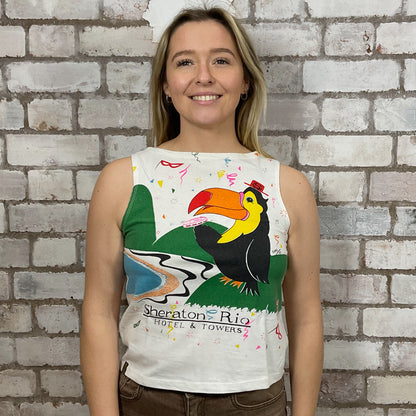 Woman wearing a colorful sleeveless top with a toucan design against a brick wall.  One-of-a-kind Rio Carnival 1993 graphic Slash Neck Unwantee tank top front view, upcycled, sustainable, made in Britain