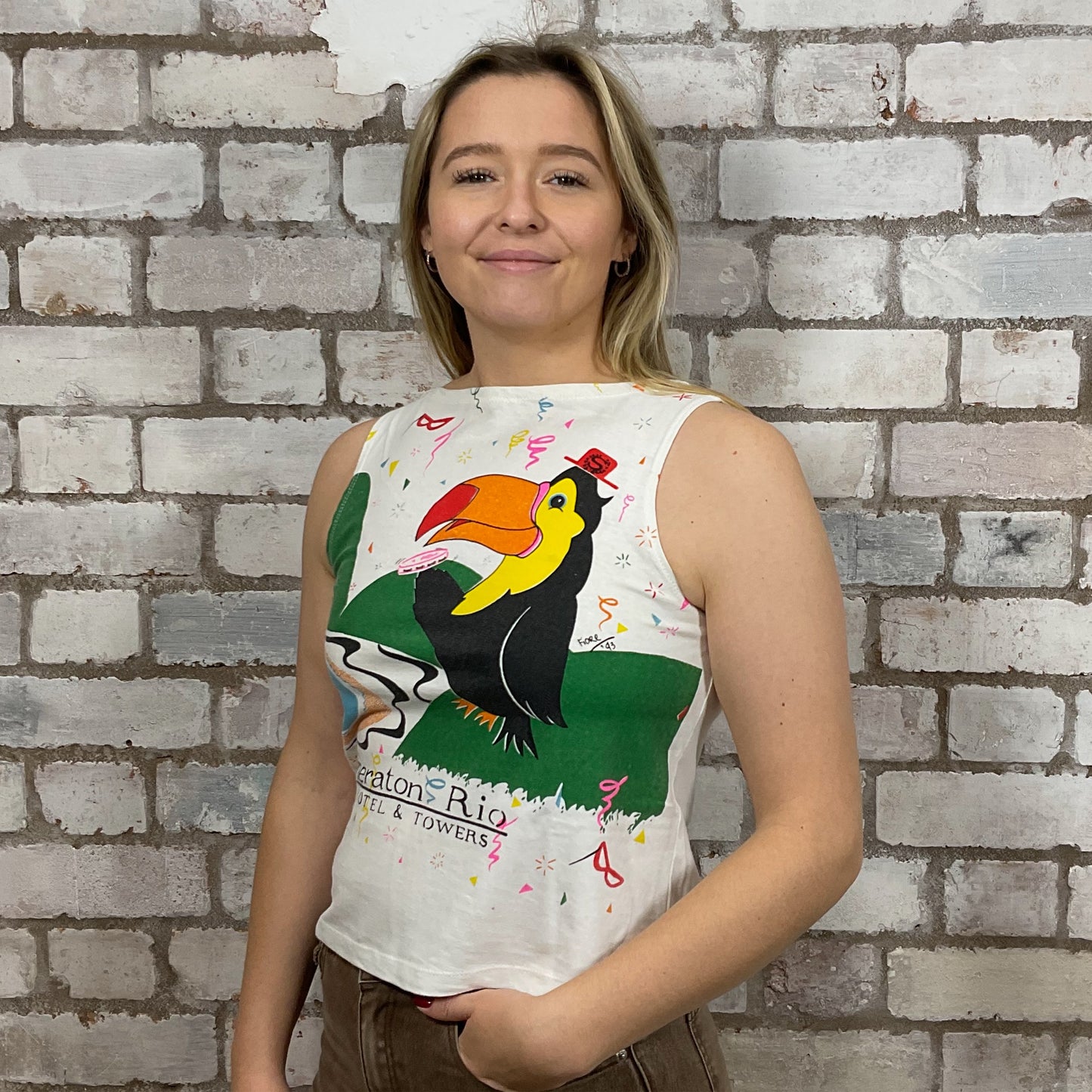 Woman wearing a sleeveless shirt with a toucan design against a brick wall.  One-of-a-kind Rio Carnival 1993 graphic Slash Neck Unwantee tank top front view, upcycled, sustainable, made in Britain