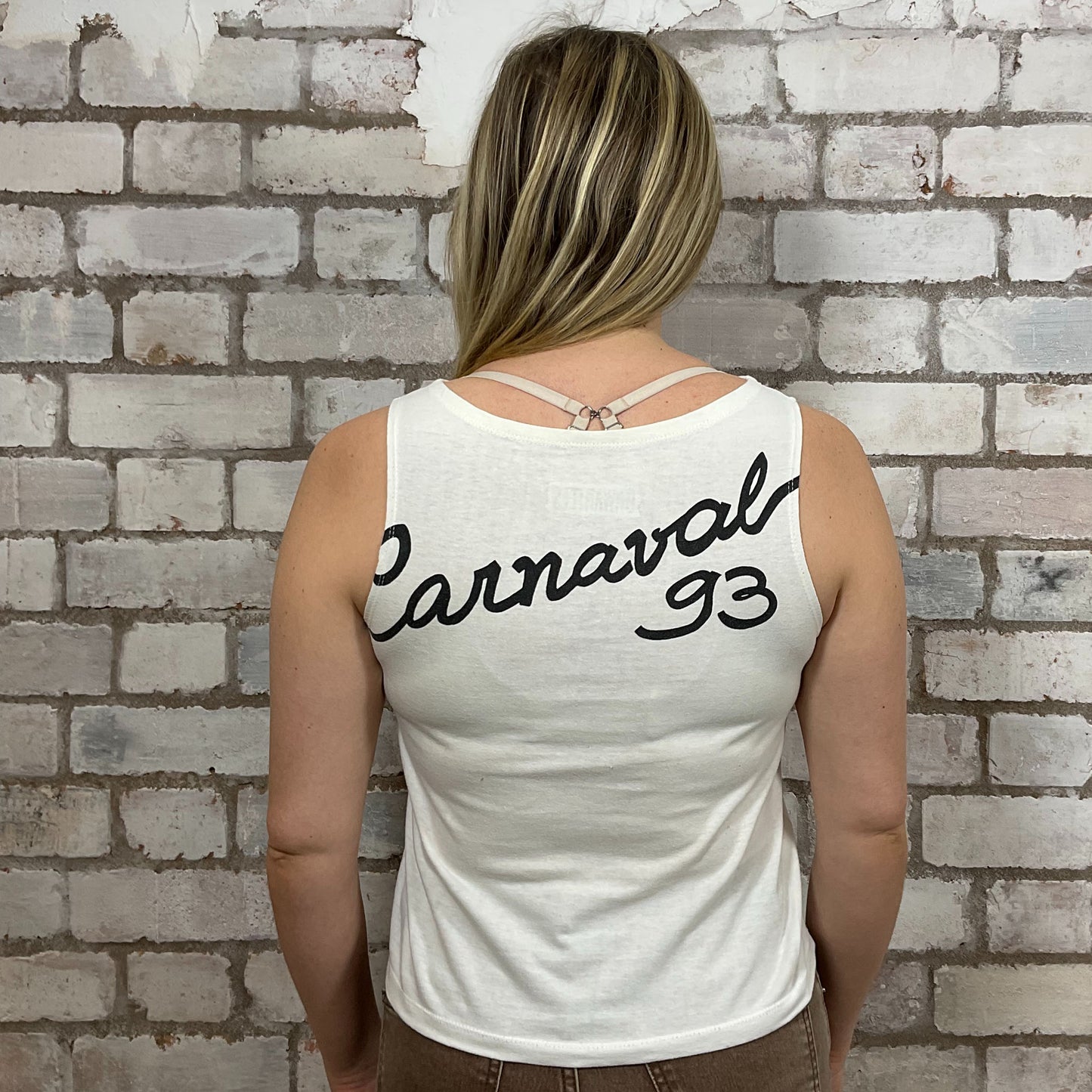Person wearing a white tank top with 'Carnaval 93' text against a brick wall.  One-of-a-kind Rio Carnival 1993 graphic Slash Neck Unwantee tank top front view, upcycled, sustainable, made in Britain