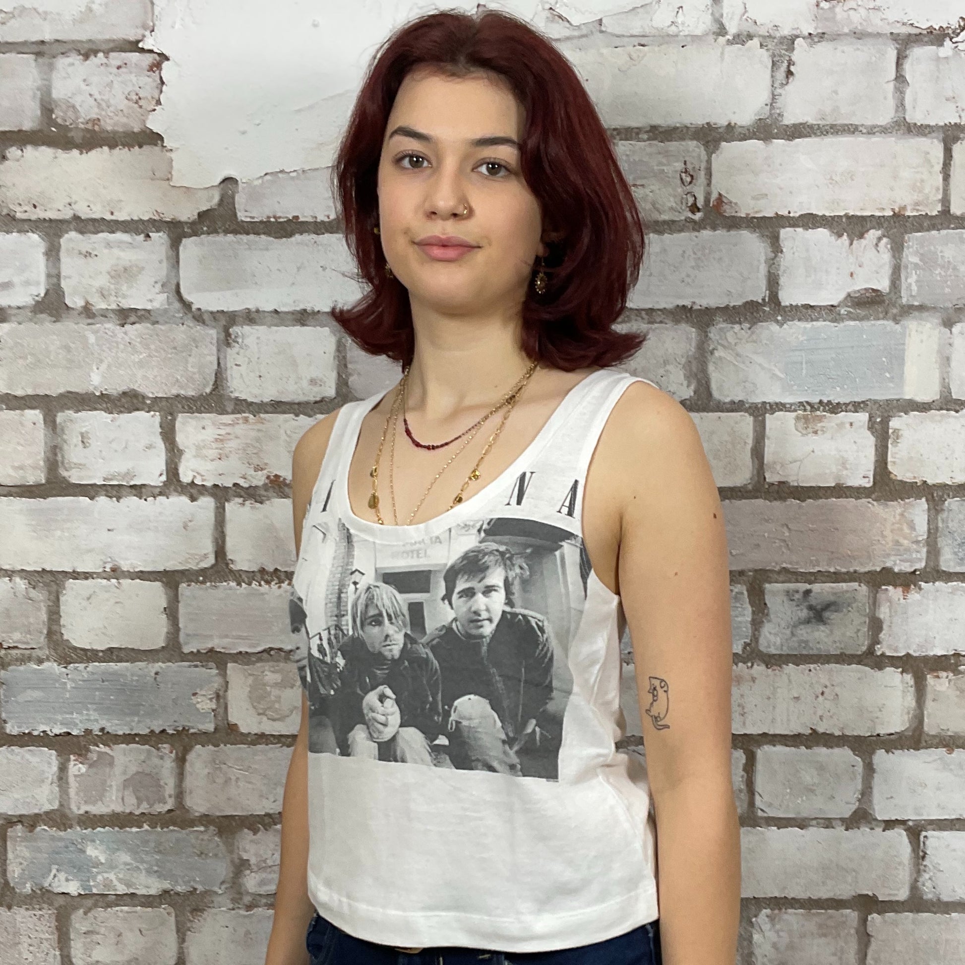 Woman wearing a tank top with a graphic design against a brick wall.  Person wearing a white tank top with Nirvana young band photo graphic design against a brick wall.