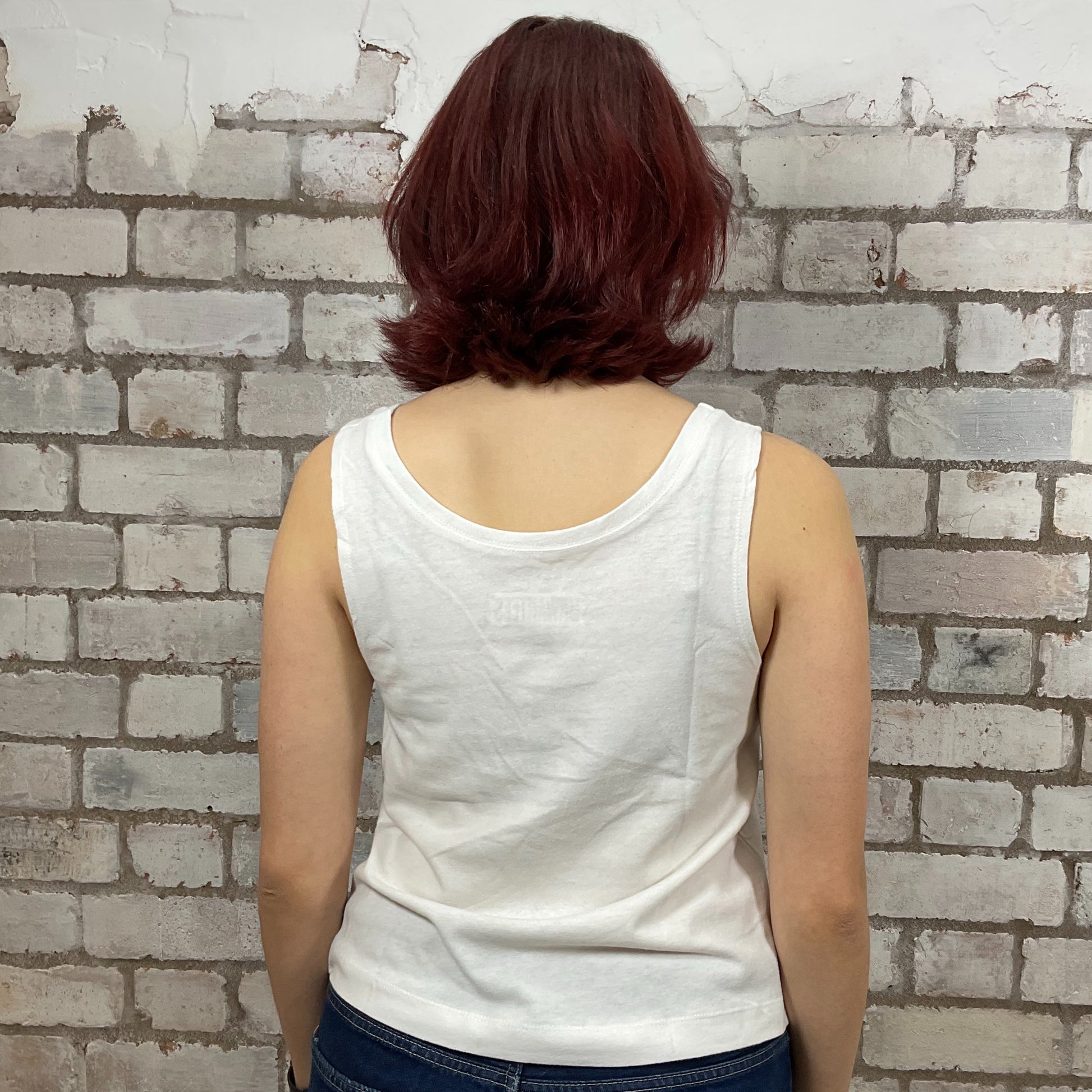 Person wearing a white tank top against a textured brick wall.  Person wearing a white tank top with Nirvana young band photo graphic design against a brick wall.