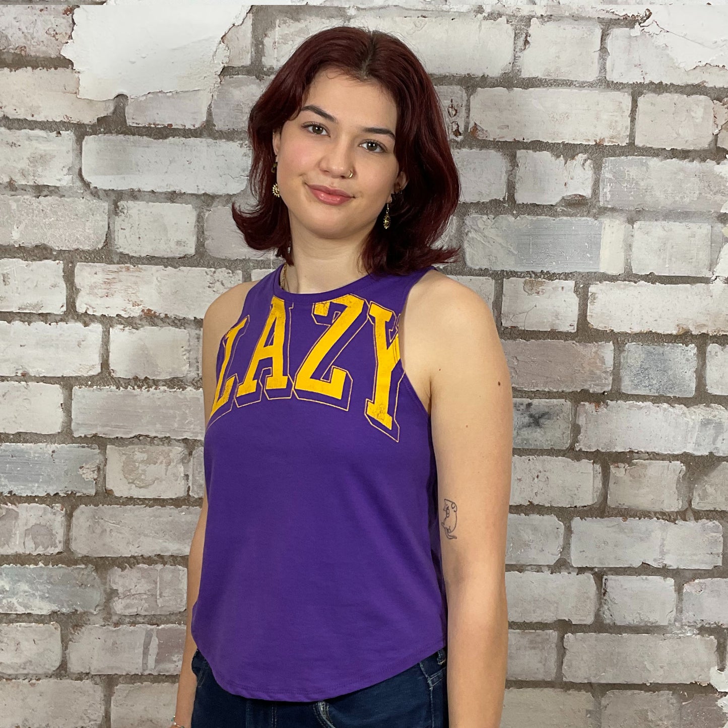 Person wearing a purple tank top with 'LAZY' printed in yellow against a brick wall.  Lazy Varsity style tshirt upcycled by unwantees in the uk, one of a kind tank tops