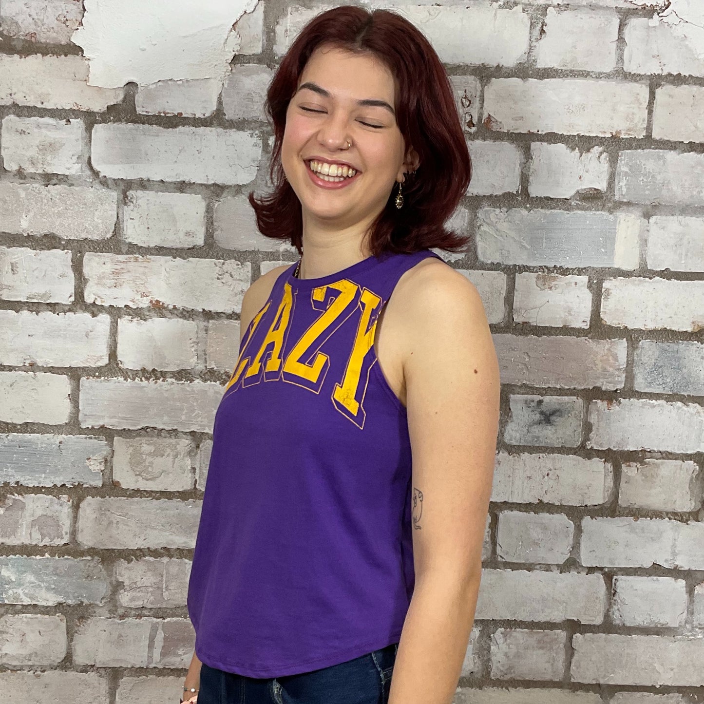 Woman wearing a purple tank top with yellow text against a brick wall.  Lazy Varsity style tshirt upcycled by unwantees in the uk, one of a kind tank tops