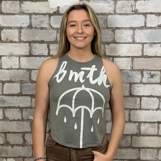 Person wearing a sleeveless shirt with 'bmth' and an umbrella graphic against a brick wall.  bmth Unwantees, Recycled clothing, sustainable fashion, athleisure wear, band tees, upcycled graphic tshirts, fashion recycled, Sustainable Fashion