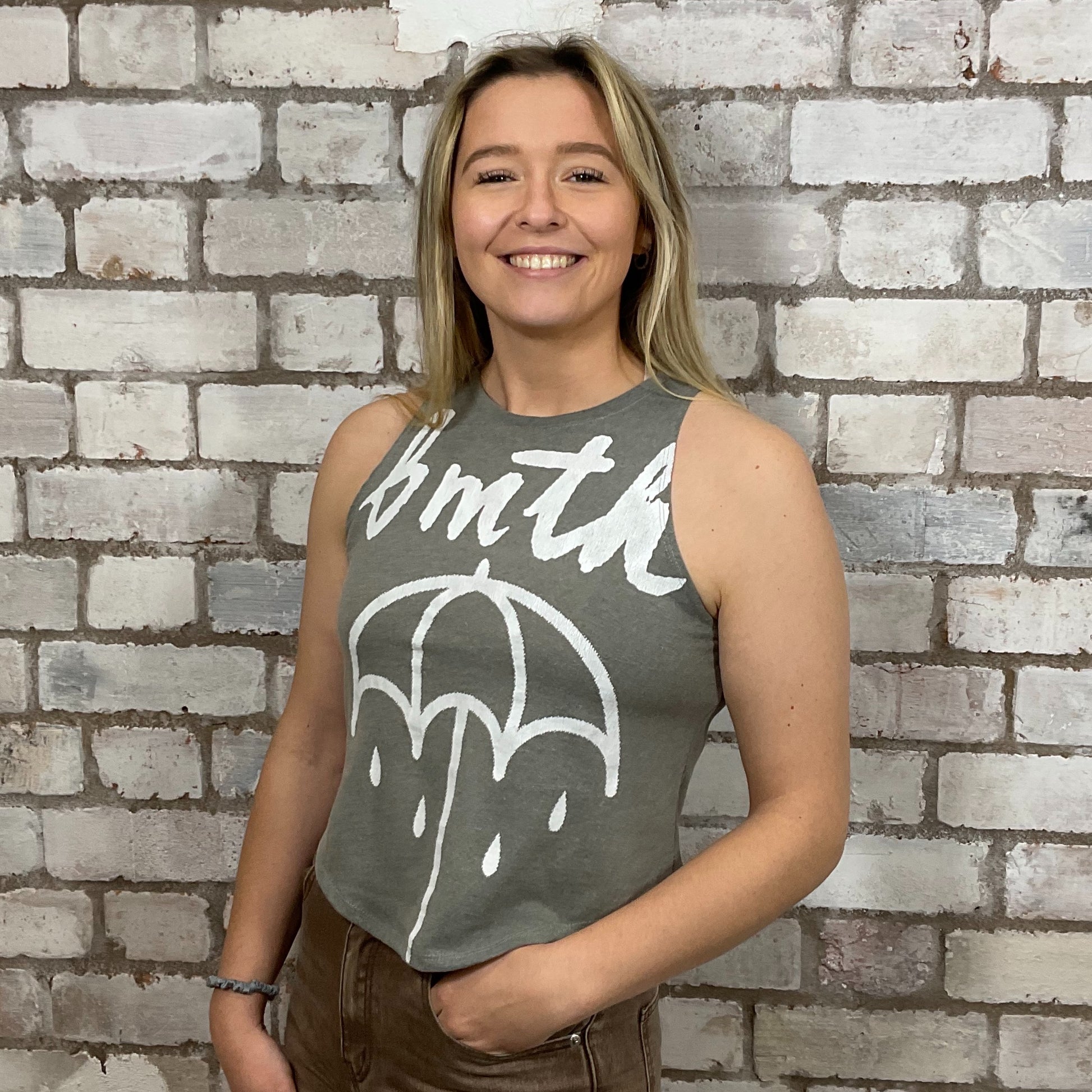 Woman wearing a sleeveless shirt with a graphic design against a brick wall.  bmth Unwantees, Recycled clothing, sustainable fashion, athleisure wear, band tees, upcycled graphic tshirts, fashion recycled, Sustainable Fashion