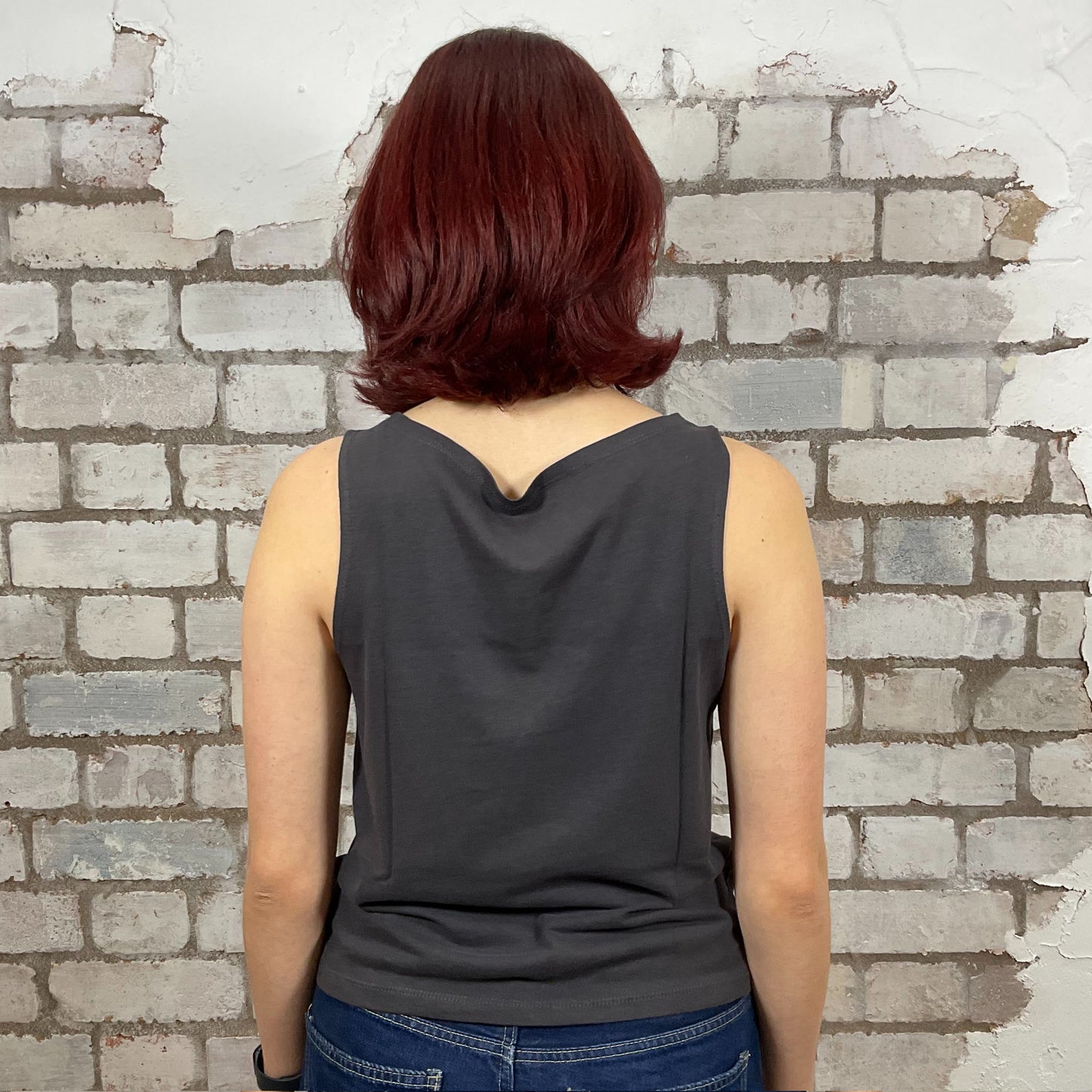 Person wearing a dark gray sleeveless top with a brick wall background.  One-of-a-kind Fleetwood Mac graphic Slash Neck Unwantee tank top front view, upcycled, sustainable, made in Britain