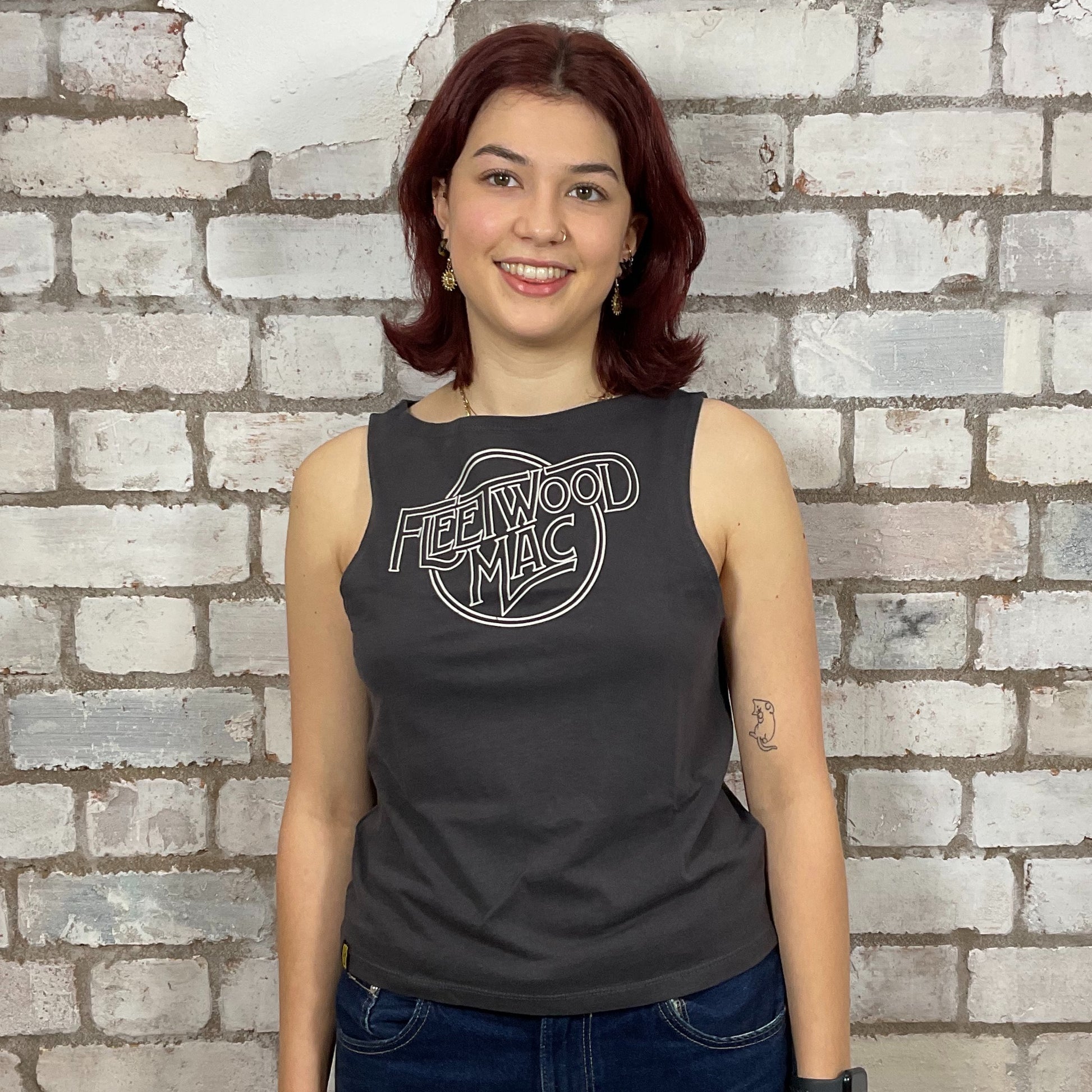 Person wearing a black sleeveless shirt with 'Fleetwood Mac' logo against a brick wall.  One-of-a-kind Fleetwood Mac graphic Slash Neck Unwantee tank top front view, upcycled, sustainable, made in Britain