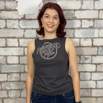 Person wearing a black sleeveless shirt with 'Fleetwood Mac' logo against a brick wall.  One-of-a-kind Fleetwood Mac graphic Slash Neck Unwantee tank top front view, upcycled, sustainable, made in Britain