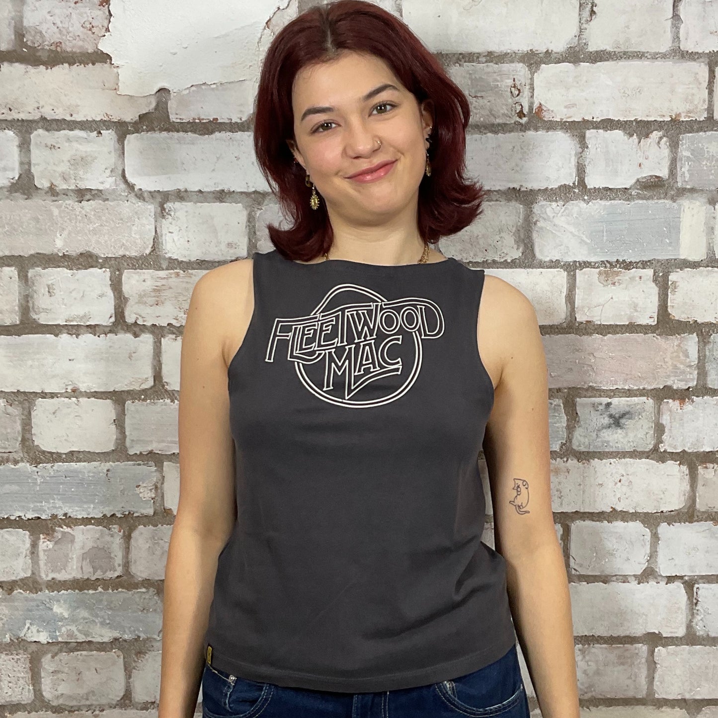 Person wearing a black sleeveless shirt with 'Fleetwood Mac' logo against a brick wall.  One-of-a-kind Fleetwood Mac graphic Slash Neck Unwantee tank top front view, upcycled, sustainable, made in Britain