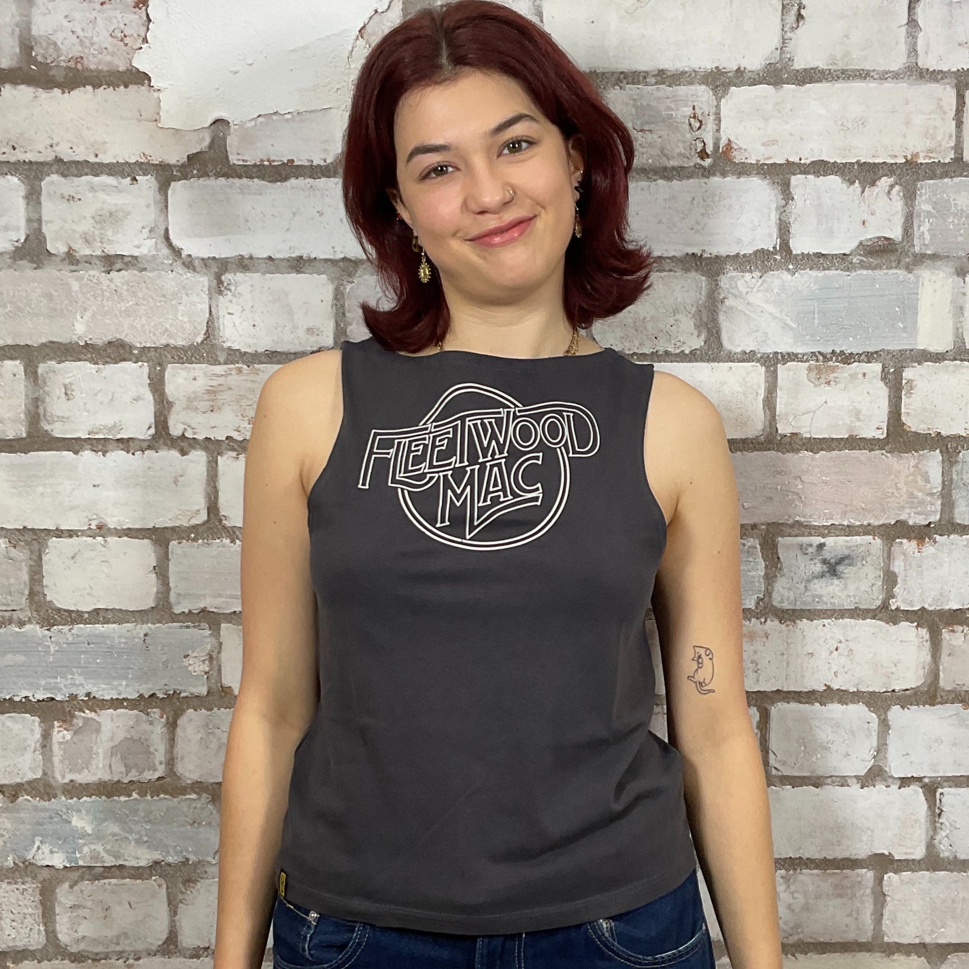 Person wearing a black sleeveless shirt with 'Fleetwood Mac' logo against a brick wall.  One-of-a-kind Fleetwood Mac graphic Slash Neck Unwantee tank top front view, upcycled, sustainable, made in Britain