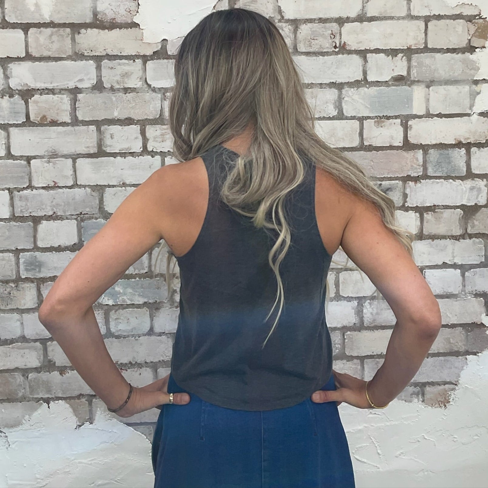 Woman standing with hands on hips against a textured white brick wall.  Rear view of racerback Unwantees