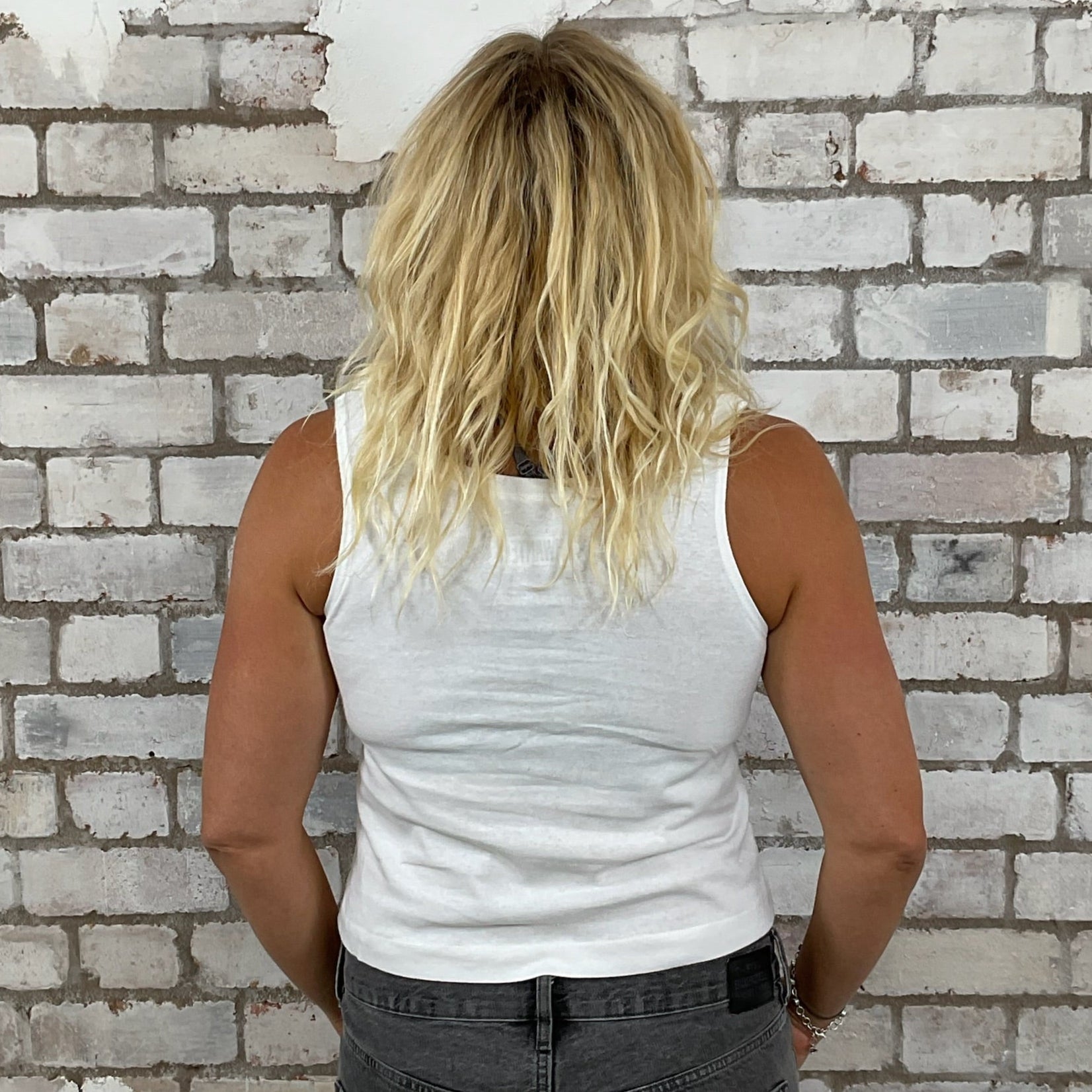 Person wearing a white tank top and dark pants standing against a textured brick wall.  Unwantees classic slash neck in white