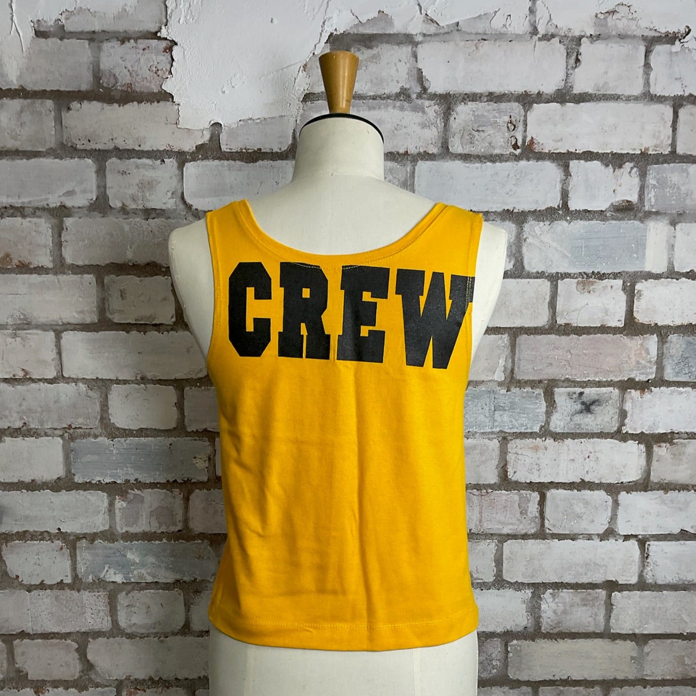 Yellow tank top with 'CREW' text on a mannequin against a brick wall.