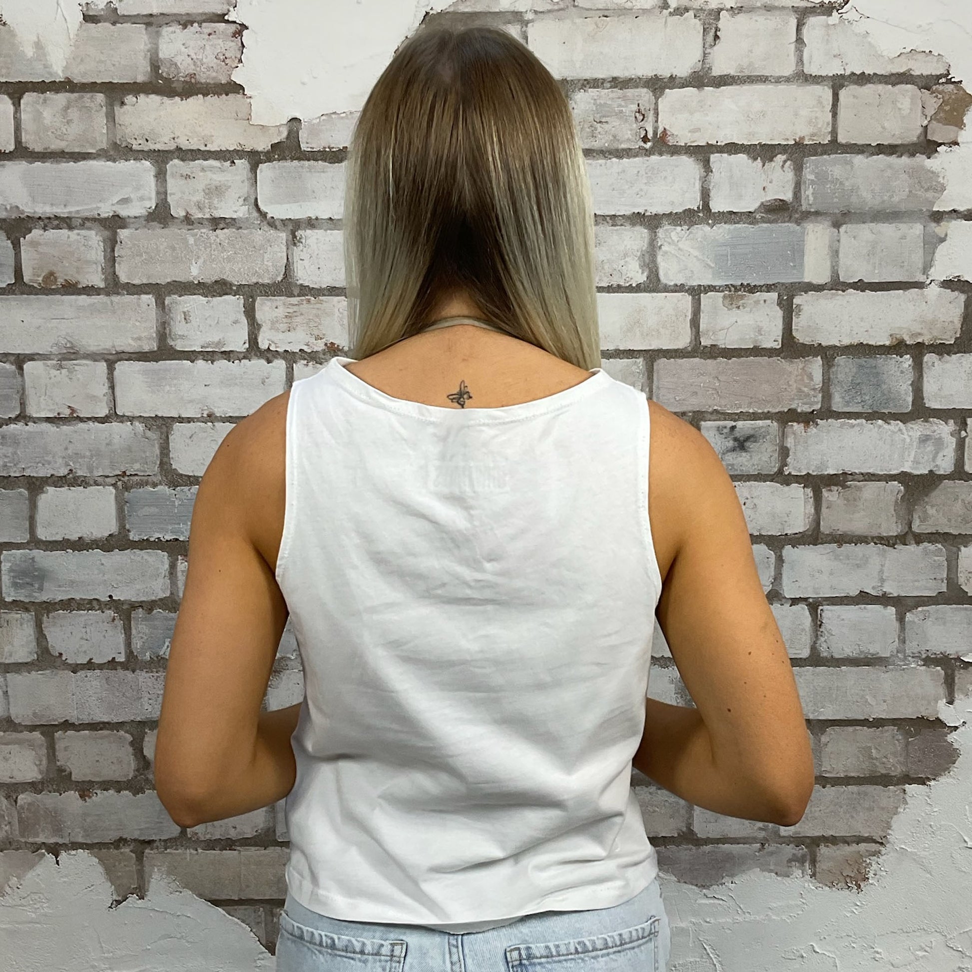 Person wearing a white tank top standing against a textured brick wall.