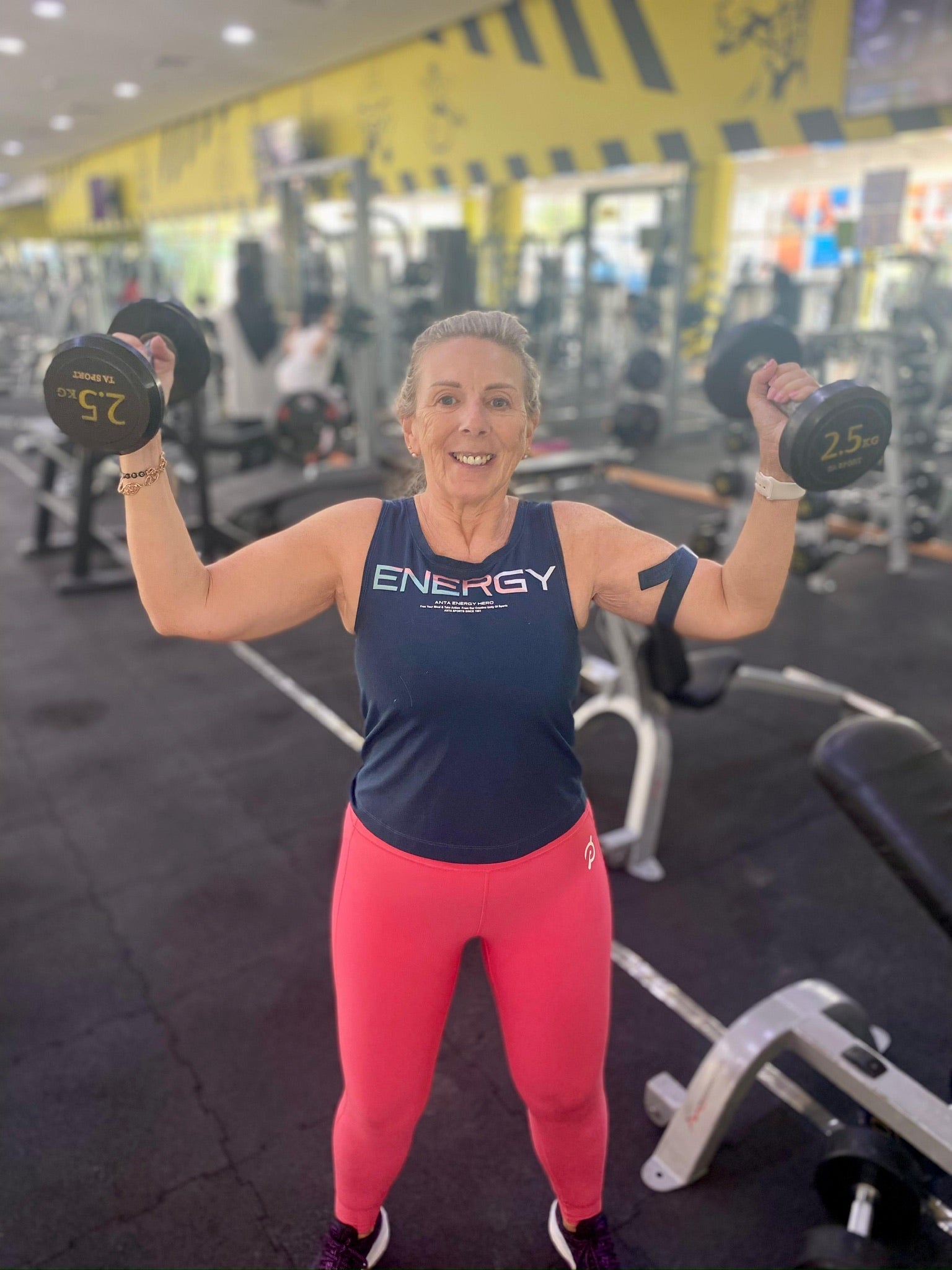 Older woman holding 2.5kg dumbbells in each hand at the gym, wearing an Unwantee top with "Energy" design, upcycled sustainable clothing